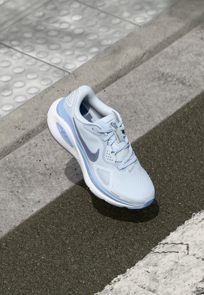 Chaussure de course Nike bleu clair avec semelle blanche reposant sur l'asphalte près d'un revêtement tactile et d'un trottoir.