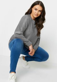 Grå långärmad topp med ribbad textur och nitar på axlarna, parat med blå jeans och vita sneakers.