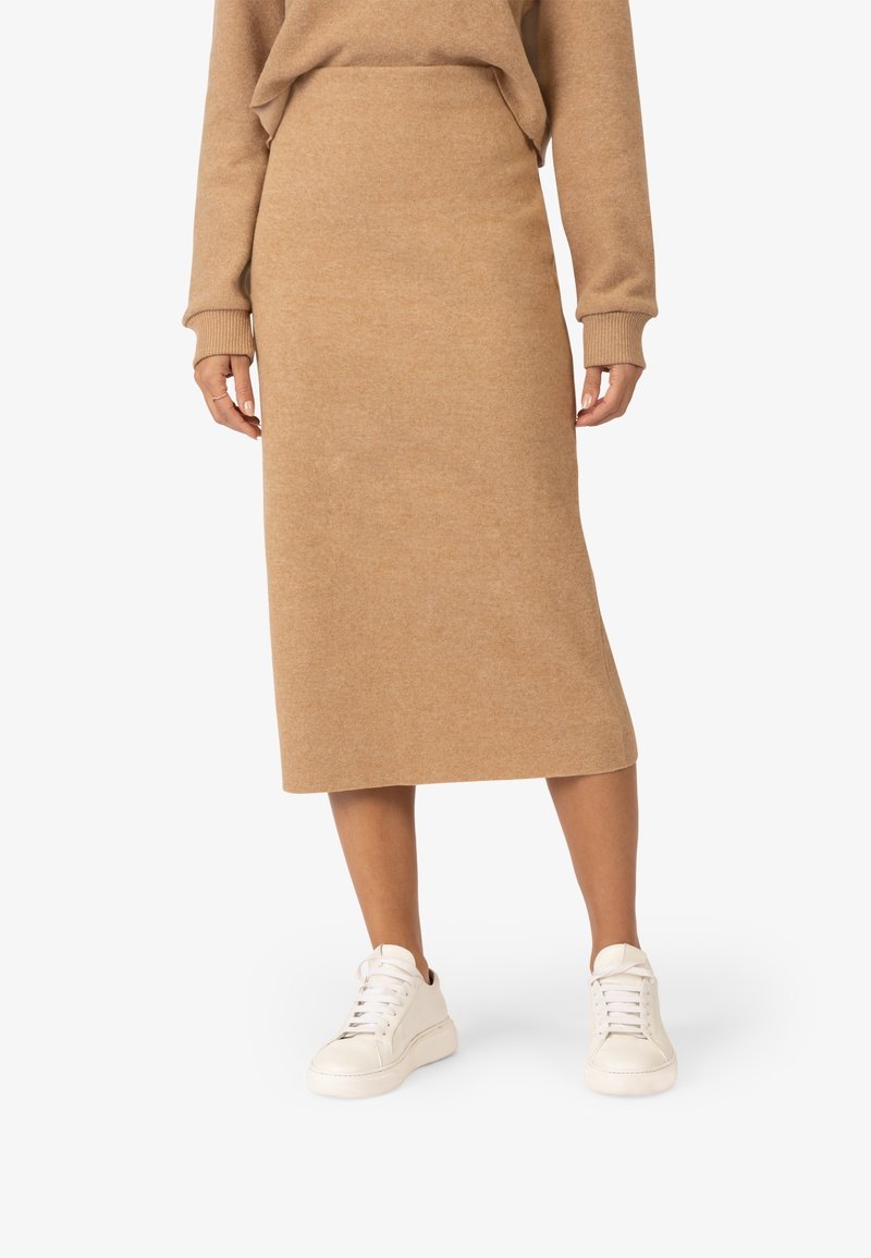 Jupe midi beige en tricot, taille ajustée, texture lisse, associée à des baskets blanches. Design simple sans embellissements ni motifs.