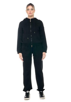 Une femme présente un sweat-shirt noir à capuche et fermeture éclair, un pantalon de jogging assorti et des baskets noires sur fond blanc.