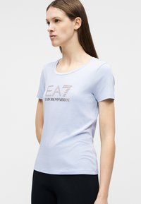 Νεαρή γυναίκα με ίσια καστανά μαλλιά φοράει ανοιχτό μπλε κοντομάνικη μπλούζα με το λογότυπο "EA7 Emporio Armani" από στρας.