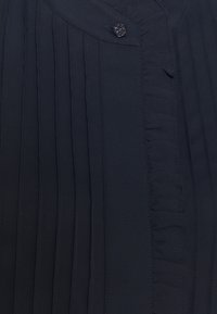 Navy pleated blouse featuring a button closure, ruffle detail along the placket, and soft-textured fabric. Simple, tailored design.