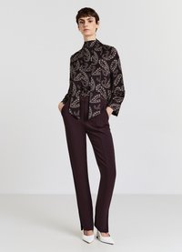 Schwarze Bluse mit Paisleymuster und hohem Kragen sowie Schleifendetail, kombiniert mit maßgeschneiderten weinroten Hosen und weißen spitzen Absätzen.