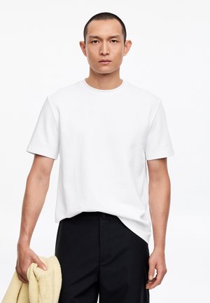 Mann trägt ein schlichtes weißes T-Shirt und schwarze Hose, hält einen gefalteten beigen Pullover in der linken Hand und steht vor einem weißen Hintergrund.