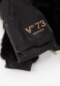 Giacca di pelle nera con bordi in pelliccia, patch con logo dorato "V°73 VENEZIA" e zip dorata, che mette in mostra un design elegante e moderno.