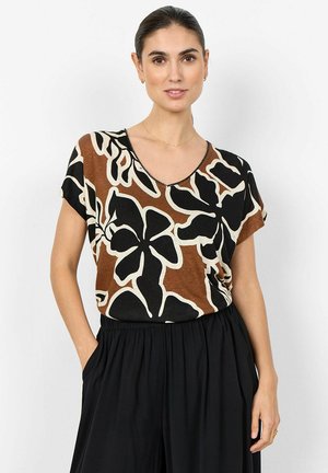 Žena s tmavými vlasmi stiahnutými dozadu má na sebe hnedo-čierno-biele tričko s kvetinovým vzorom s krátkym rukávom a čierne nohavice s vysokým pásom, stojí s rukami v vreckách.