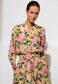 Bloemige jurk met roze, gele en groene patronen. Lichtgewicht stof, lange mouwen, een v-hals en een strik in de taille. Geplooide rok.