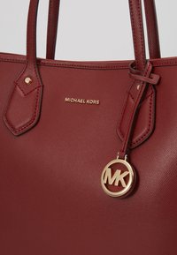 Sac fourre-tout en cuir rouge à finition texturée, avec un pendentif logo doré et un détail de marque sur le devant. Deux poignées supérieures.