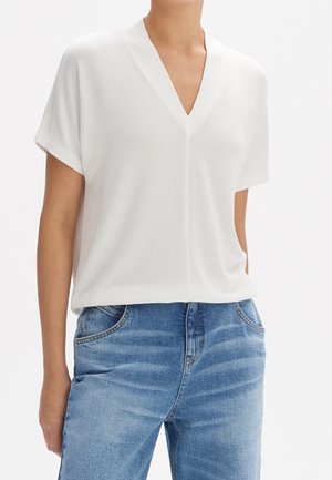 Femme portant un haut blanc à manches courtes et col en V, associé à un jean bleu clair délavé sur un fond blanc uni.