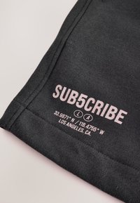 Mørkegrå t-shirt med lyserød trykt tekst, herunder "SUB5CRIBE" og koordinater for Los Angeles, CA. Blød tekstur og rund hals.