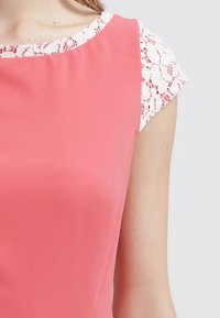 Kobieta w koralowo-różowej sukience z białymi koronkowymi krótkimi rękawami oraz koronkowym wykończeniem dekoltu, pokazana od ramienia do środka torsu.