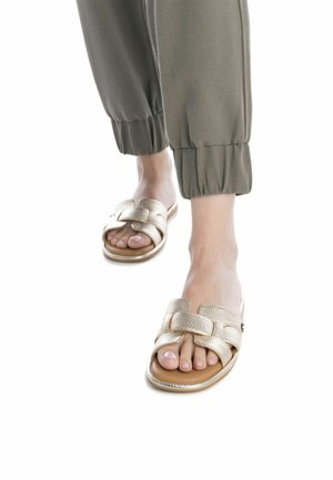 Las sandalias deslizantes con textura dorada presentan un diseño de tira ancha entrelazada y una suela plana color marrón claro, combinadas con pantalones deportivos de color verde oliva.