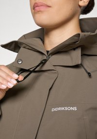 Pruun veekindel jope lukuga ees, reguleeritava nööriga kapuutsiga ja logoga. Omadusteks on sile, tekstuuriga viimistlus ja mustad riistvaradetailid.