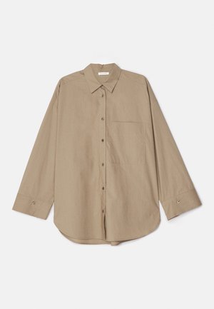 Henkilö, jolla on beige midimekko ja ylisuur tan-nappipaita sekä ruskeat punotut sandaalit, seisoo harmaalla lattialla.