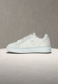 Λευκό sneaker χαμηλού ύψους με λευκά κορδόνια, λογότυπο χαραγμένο στο πλάι και παχιά λευκή σόλα, εμφανιζόμενο σε ουδέτερο φόντο.