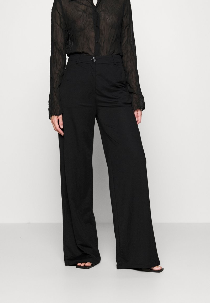Femme portant un chemisier noir transparent à manches longues et un pantalon noir taille haute à jambes larges avec des sandales noires ouvertes aux orteils.