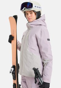 Veste de ski en violet clair et beige, dotée d'une capuche, de poches zippées et d'un logo Roxy. Skis avec base cuivrée tenus dans la main gauche.