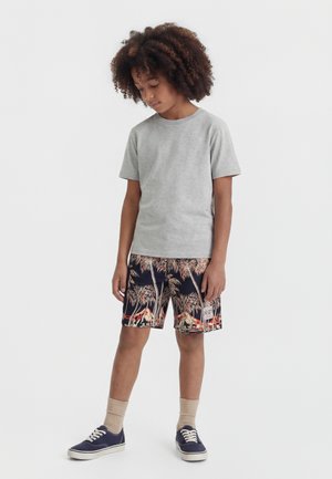 Barn med krøllete hår står og ser ned, iført en grå t-skjorte, shorts med tropisk mønster, beige sokker og marineblå joggesko på hvit bakgrunn.