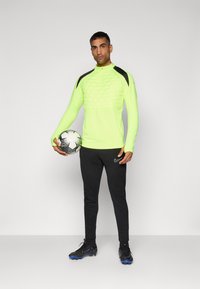 Neon-gelbes Langarm-Sportoberteil mit strukturiertem, hexagonalem Muster auf der Brust, schwarzen Schulterakzenten, kombiniert mit schwarzen Hosen und einem Fußball.