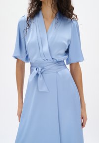 Helles blaues Kleid mit einem Wickeldesign, kurzen Ärmeln, Gürteldetail und strukturiertem Stoff. Der Rock ist fließend und knielang.