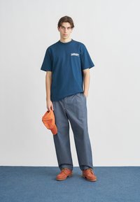 Μπλε σκούρο βαμβακερό μπλουζάκι με το λογότυπο "CARHARTT", γκρι παντελόνι φαρδιάς γραμμής, πορτοκαλί καπέλο στο χέρι και καφέ δερμάτινα παπούτσια με κορδόνια, στέκεται πάνω σε μπλε χαλί.