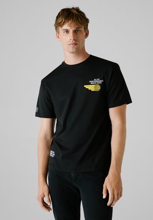 Zwarte katoenen T-shirt met korte mouwen, voorzien van een gele grafisch ontwerp met tekst en vleugelmotief op de voorkant. Effen achterkant.