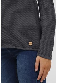 Dark gray textured sweater with a knitted pattern and long sleeves. Features a wooden logo accent near the hem. Paired with denim jeans.