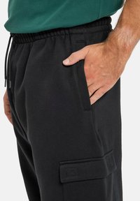 Schwarze Sweathosen mit einem elastischen Bund, Seitentaschen und einer Cargo-Tasche mit genähten Akzenten. Das Material wirkt weich und strukturiert.