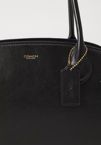 Schwarze Lederhandtasche mit goldenem Logo, oberen Griffen und einem rechteckigen Anhänger. Glatte Textur, abgerundete Form und auffällige Steppdetails.