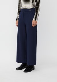 Marineblauwe wijde pijpenbroek met een hoge taille, knoopdetail en plooien aan de voorkant, gemaakt van een gladde stof, gecombineerd met zwarte schoenen.