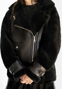 Sort faux fur- og læderjakke med bred krave, lynlås lukning og teksturerede manchetter med dekorativt accent.