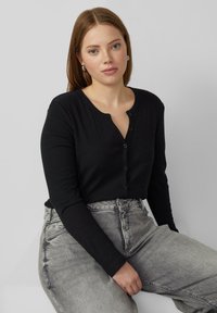 Zwart, geribd shirt met lange mouwen, een V-hals en een knoopsluiting met vijf knopen, gecombineerd met grijze spijkerbroek. Zachte textuur en aansluitend ontwerp.