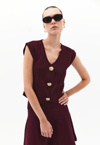 Burgunderfarbene Strickbluse mit V-Ausschnitt, kurzen Ärmeln, drei goldfarbenen Knöpfen und seitlichen Nahtdetails. Getragen mit passendem Rock.