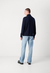 Navy Zip-Jacke mit hohem Kragen, kombiniert mit hellblauen Schlagjeans und schwarzen klobigen Schuhen, von hinten betrachtet.