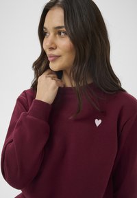 Vínová mikina s žebrovanými manžetami a límečkem. Na hrudi má malý bílý srdíčkový znak. Měkký materiál a volný střih.