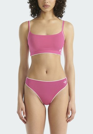 Woman wearing a pink sports bra and matching high-cut underwear with white trim and small Adidas logos on both pieces.
