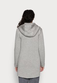 Cappotto grigio a maniche lunghe con cappuccio, realizzato in tessuto morbido. Presenta un orlo curvo e polsini aderenti. Design semplice, senza motivi o elementi metallici visibili.