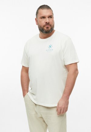 Homme avec une barbe et les cheveux attachés, portant un T-shirt blanc avec un logo bleu et un pantalon beige, debout avec une main dans la poche.