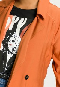 Pomarańczowa lekka kurtka z klapami, pojedynczym czarnym guzikiem, nałożona na czarny t-shirt z graficzną ilustracją.