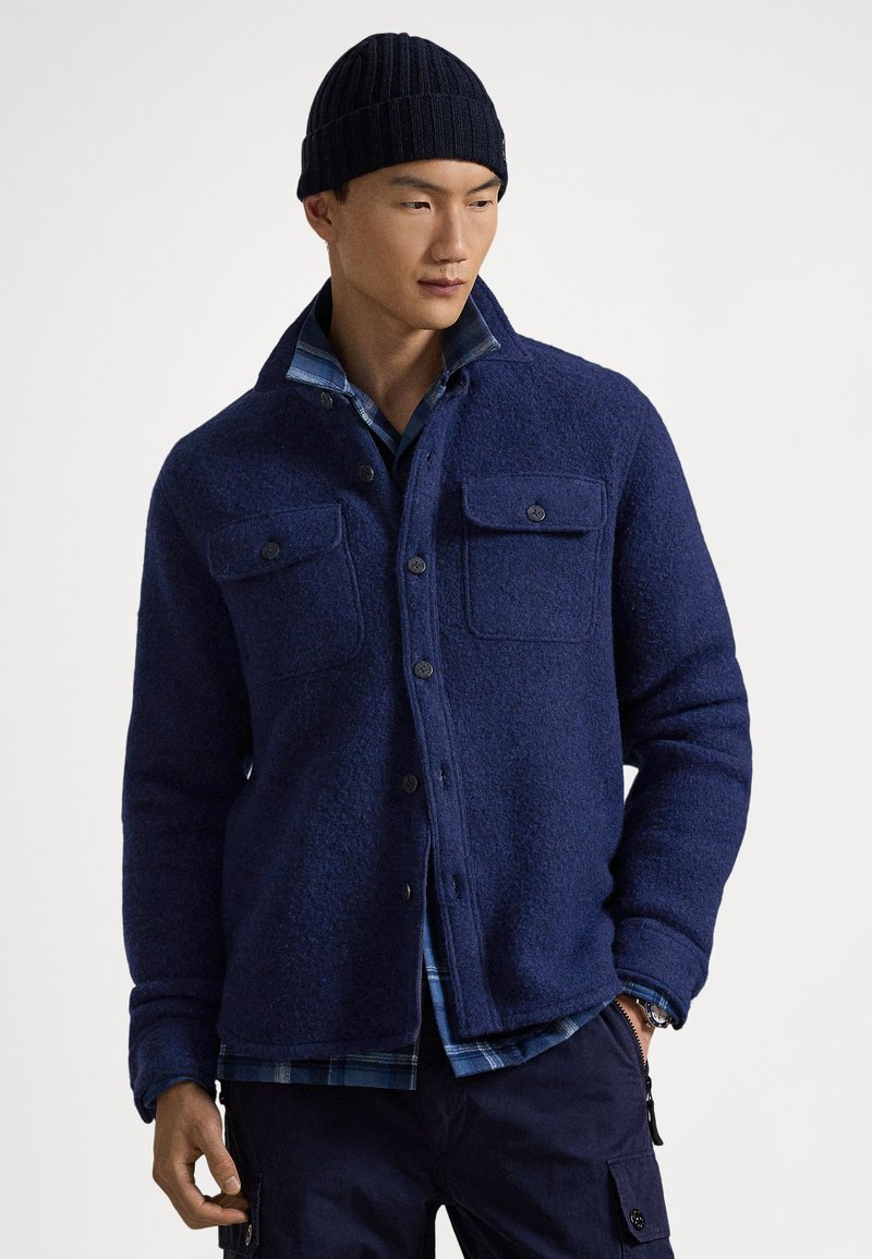 Marineblaue Wolljacke mit geknöpftem Vorderteil, zwei Brusttaschen und einem darunter getragenen mehrlagigen karierten Hemd. Abgerundet mit einer gerippten Strickmütze.