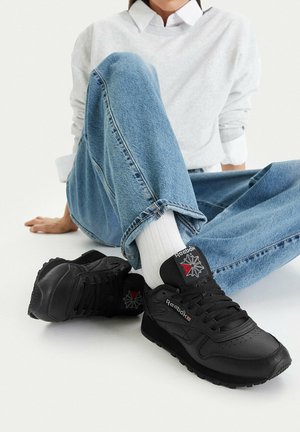 Osoba sedící se zkříženýma nohama, oblečená v světlých modrých džínách, bílých ponožkách a černých teniskách Reebok s červeným a bílým logem na bílém pozadí.