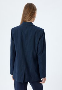 Blazer blu navy a righe con spalle strutturate, spacco singolo sul retro e rever classici. Tessuto liscio con una finitura professionale.