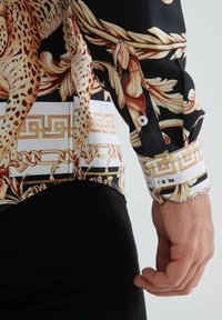 Chemise à manches longues présentant un motif noir et doré avec des motifs animaliers, une coupe ajustée et des boutons décoratifs aux poignets et à l'ourlet.