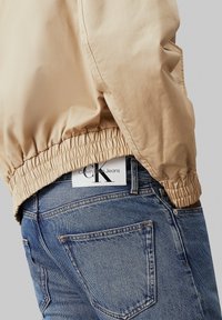 Beige bomberjacka med elastisk nederkant. Buren över blå denimjeans med en bakficketikett från Calvin Klein Jeans.