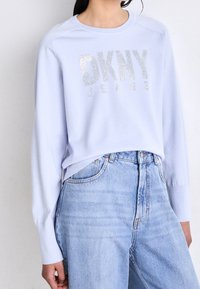 Ljusblå sweatshirt med "DKNY JEANS" i silverglittrande bokstäver. Långa ärmar, rundad halsringning, avslappnad passform. Kombinerad med högmidjade jeans.