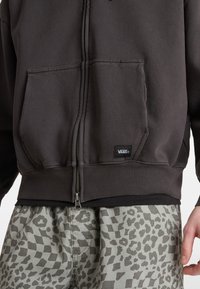 Dunkelgrauer Zip-Hoodie mit einer Fronttasche und schwarzem Marken-Tag, kombiniert mit hellgrauen Shorts im Leopardenmuster.