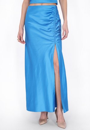 Kobieta w jasnoniebieskiej, satynowej maxi spódnicy z marszczeniami i wysokim rozcięciem, zestawiona z beżowymi szpilkami z zaostrzonym noskiem.