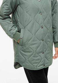 Puteruotas žalias švarkas su lygia išorine dalimi, apvaliais sidabriniais spaustukais, šoninėmis kišenėmis ir audinio tekstūra su bangelių raštu.