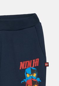 Tamno plave pamučne trenirke s grafikom dva maskirana ninje u plavoj i crvenoj boji te natpisom "NINJA" u jarko crvenoj. Elastična stražnja traka.