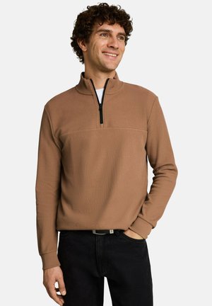Mann mit lockigem Haar, der einen braunen Pullover mit Viertel-Reißverschluss und schwarze Hose trägt, lächelt und schaut zur Seite, wobei eine Hand in der Tasche ist.